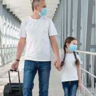 Père tenant la main de sa jeune fille portant des masques et tirant une valise